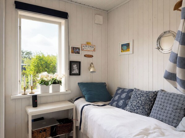A bedroom with a bed, a window with white curtains, a bedside table with a lamp, and a wall-mounted mirror.