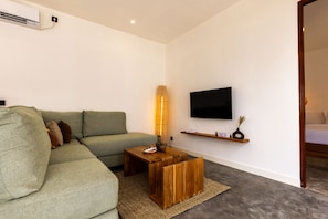 Una sala moderna con un sofá verde, una mesa de centro de madera, un televisor de pantalla plana montado en la pared y un pequeño estante de madera con objetos decorativos.