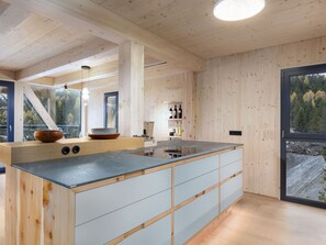 A modern kitchen with a central island, wooden cabinetry, and a view of a forest through a large window. - 03_NTH032_Deluxe Chalet mit Sauna und Hot Tub by Interhome (Großveitsch)