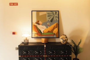 Una habitación con un cuadro enmarcado de una persona relajándose, una mesita de noche con una lámpara, un jarrón decorativo y un letrero de salida de incendios.