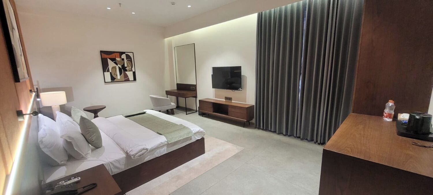 A modern hotel room with a bed, bedside table, lamp, TV, and a large window with curtains.