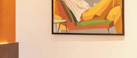 A hotel room with a bed, a chair, a small table with a glass and a green container, and a painting on the wall.