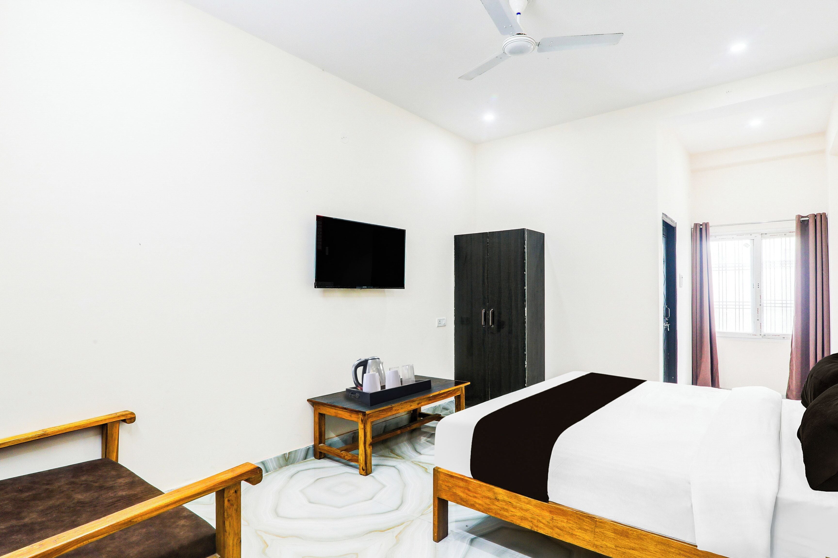 A hotel room with a bed, a TV mounted on the wall, a wardrobe, a small table with a kettle, and a window with curtains.