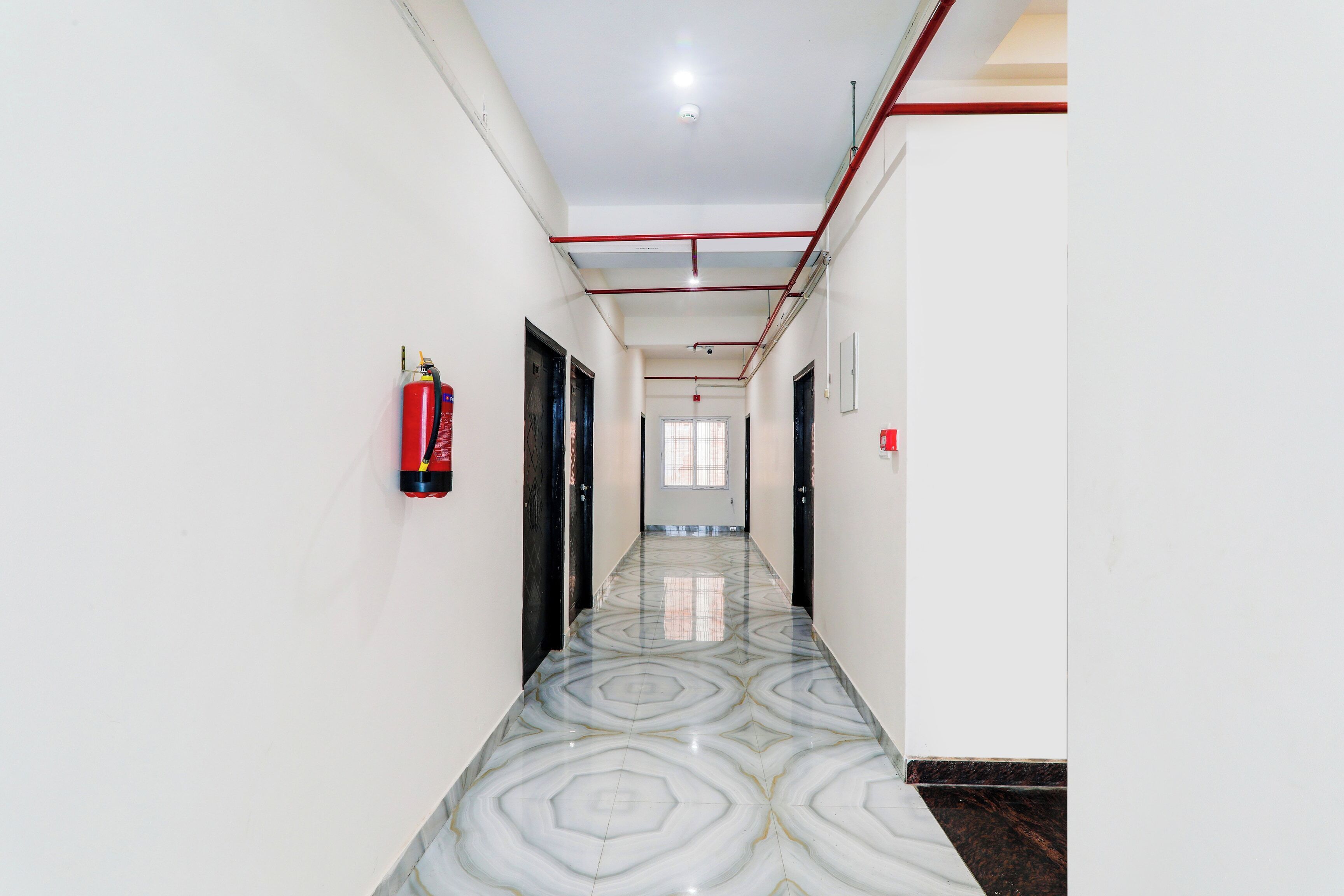 A corridor with a patterned floor, fire extinguishers, and a ceiling with visible pipes.