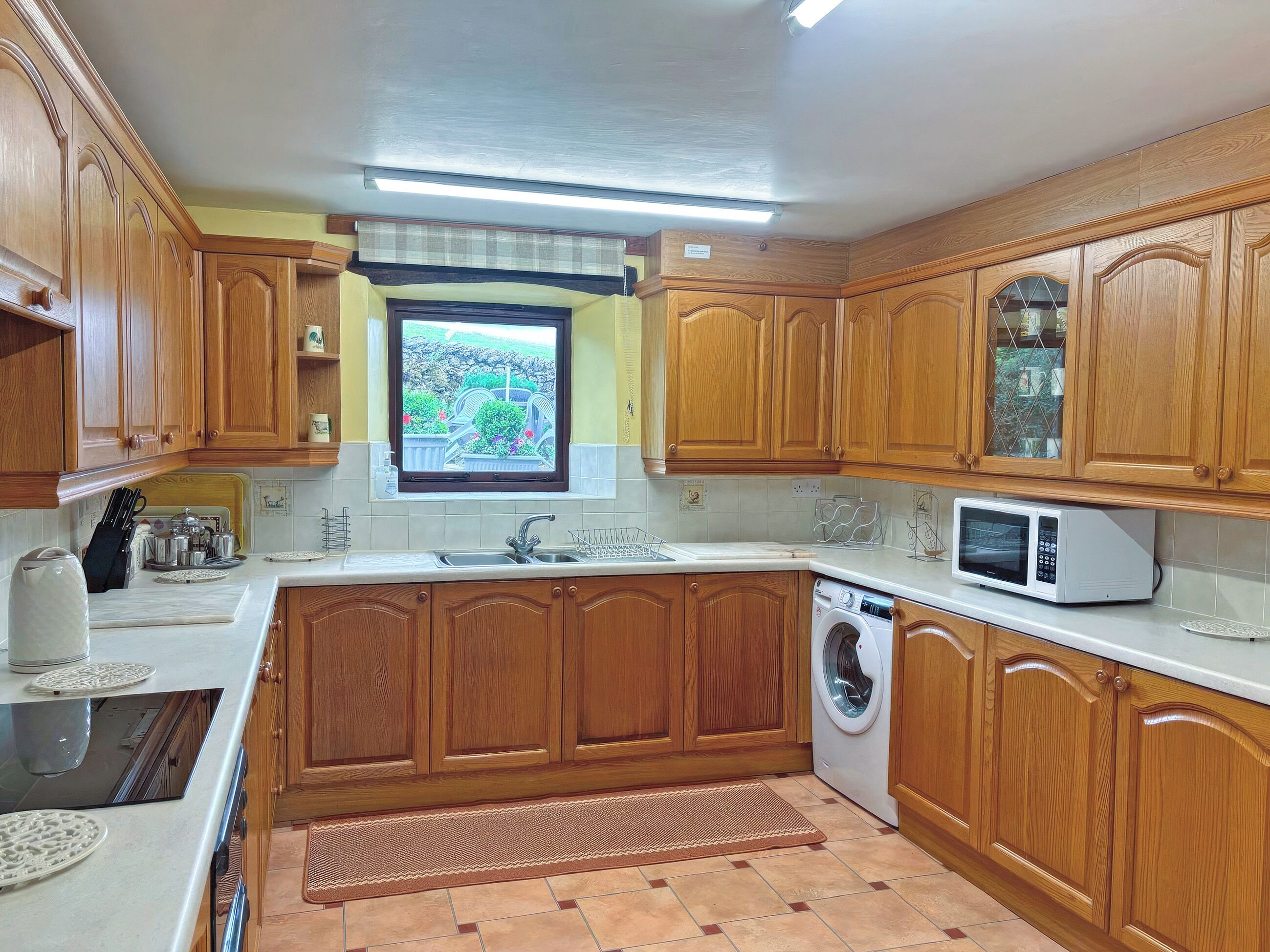 A kitchen with wooden cabinets, a built-in microwave, a washing machine, and a window with a view of greenery.
