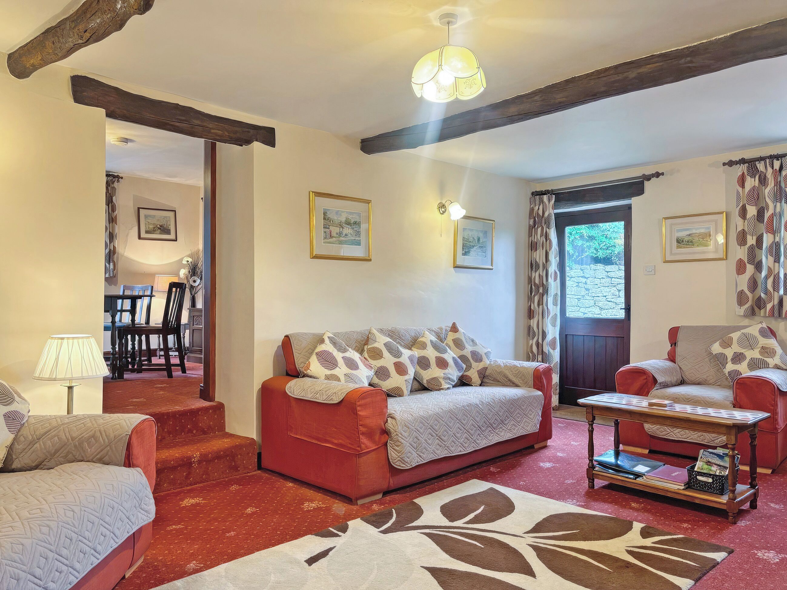 A cozy living room with a red sofa, a coffee table, and a patterned rug. There are framed pictures on the walls and a door leading outside.
