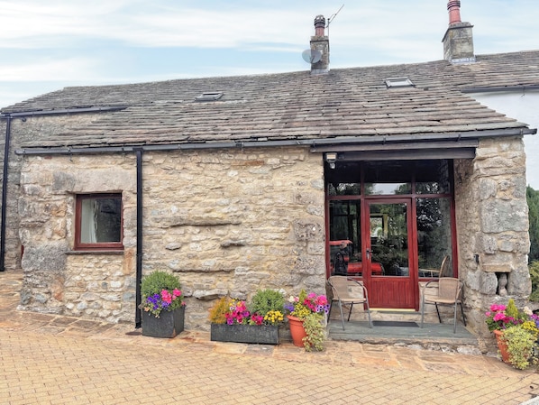 A stone building with a tiled roof, a small window, and a door leading to an outdoor seating area with chairs and potted plants. - 2 steps to entrance. (Settle)
