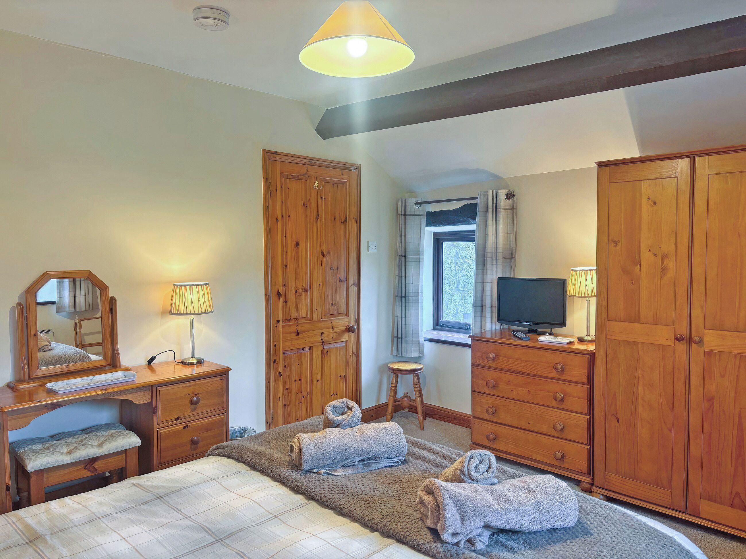 A bedroom with a bed, wooden wardrobe, a desk with a mirror, a television, and a window with curtains.