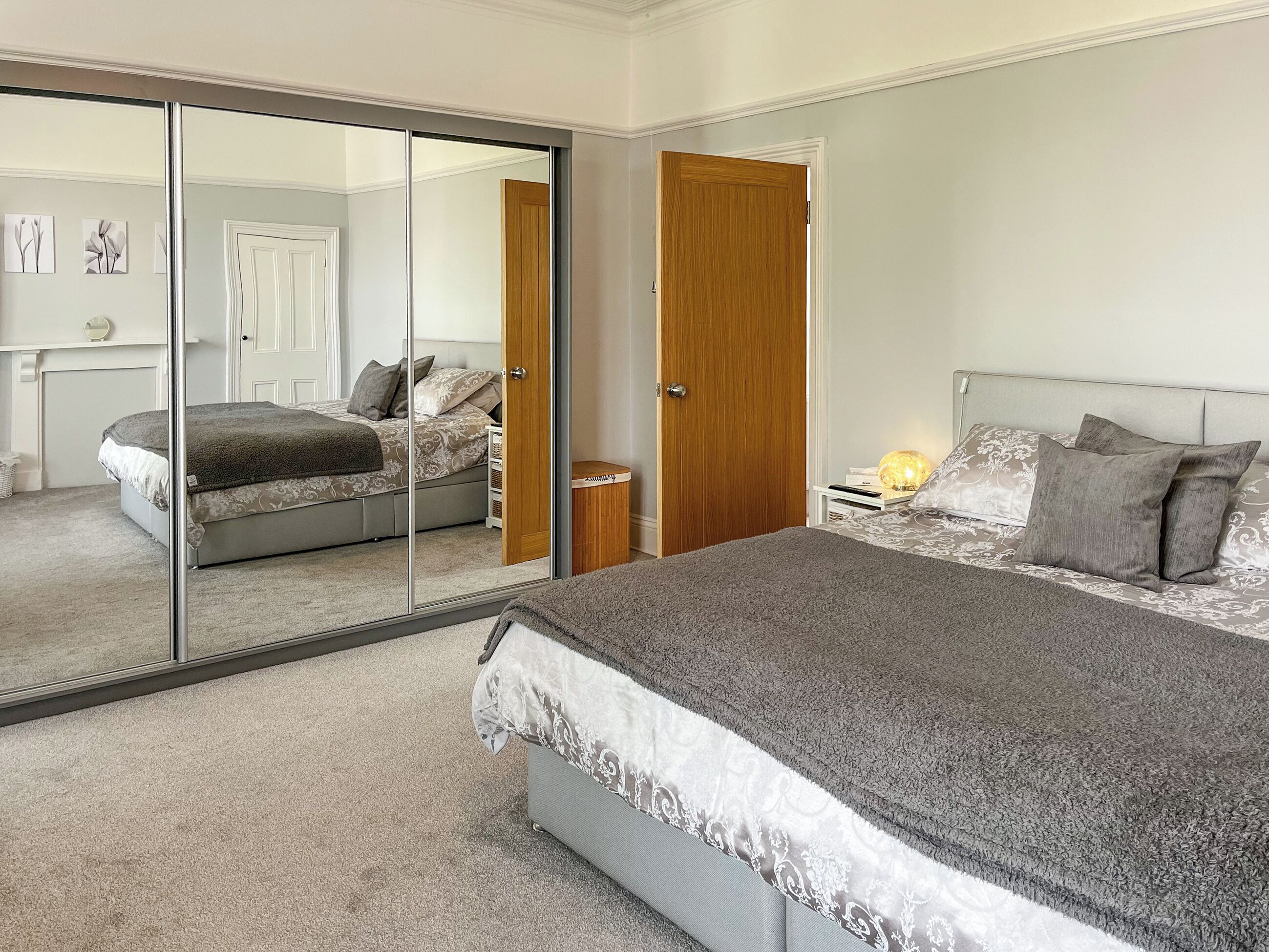 A bedroom with a large bed, a bedside table with a lamp, a wardrobe, and a sliding glass door leading to another room.
