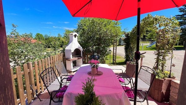 Een terras buiten met een barbecue, stoelen en een tafel onder een rode parasol.
