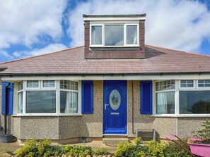 Ein zweigeschossiges Haus mit einer blauen Haustür, einem kleinen Balkon und einem Garten mit Pflanzen und einer Schubkarre.
