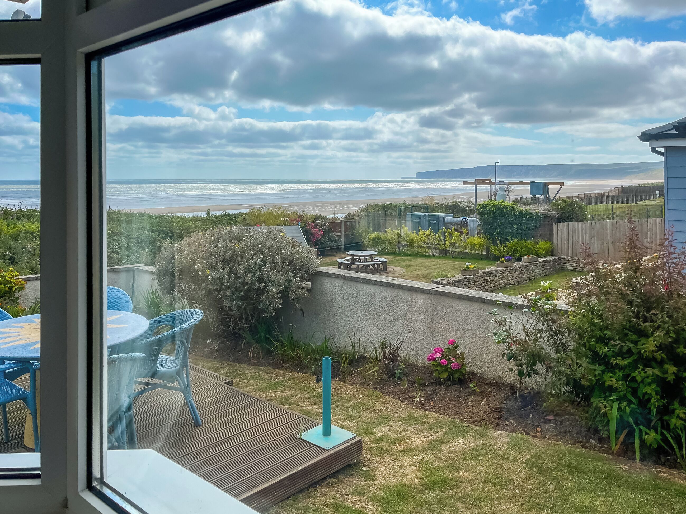 A view from a window overlooking a garden with a patio area, a wooden deck, and a view of the ocean.