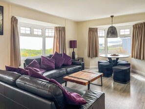 Ein modernes Wohnzimmer mit einer Ledersofa, einem Couchtisch und Hocker. Große Fenster mit Vorhängen, eine Lampe und ein Blick ins Freie sind zu sehen.