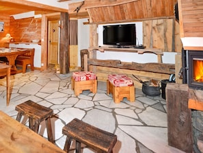 Ein rustikaler Innenraum mit Steinboden, Holzmöbeln, einem an einem Holzbalken befestigten Fernseher und einem Kamin.