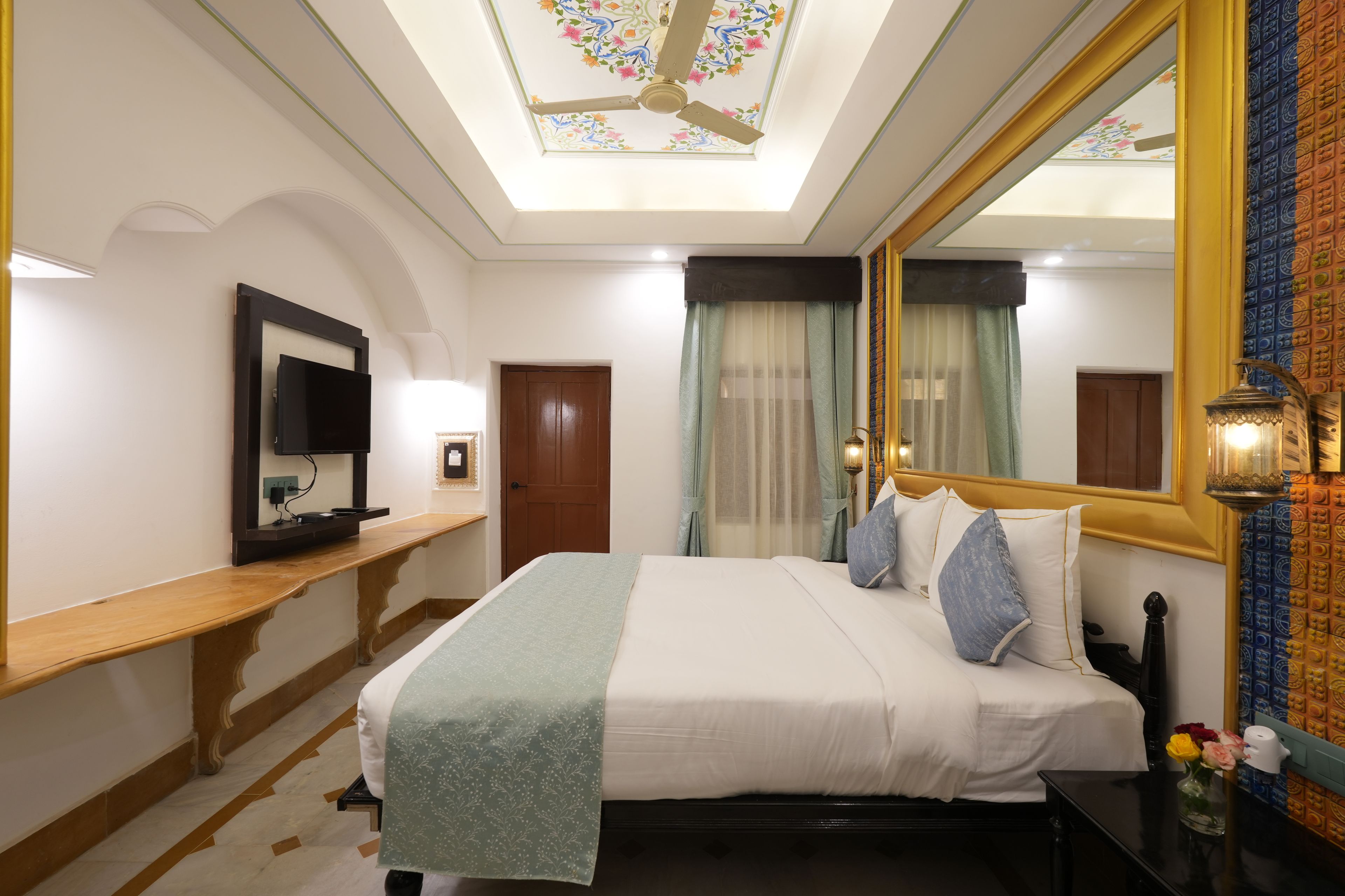 A hotel room with a bed, a bedside table, a mirror, a ceiling fan, and a window with curtains.