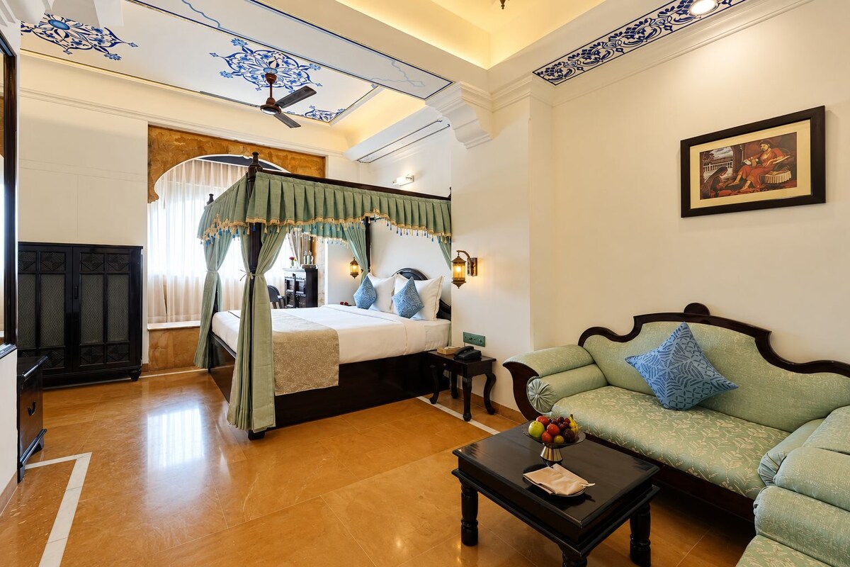 Una habitación de hotel con cama con dosel, una zona de estar con sofá, una mesa de centro y un televisor montado en la pared.