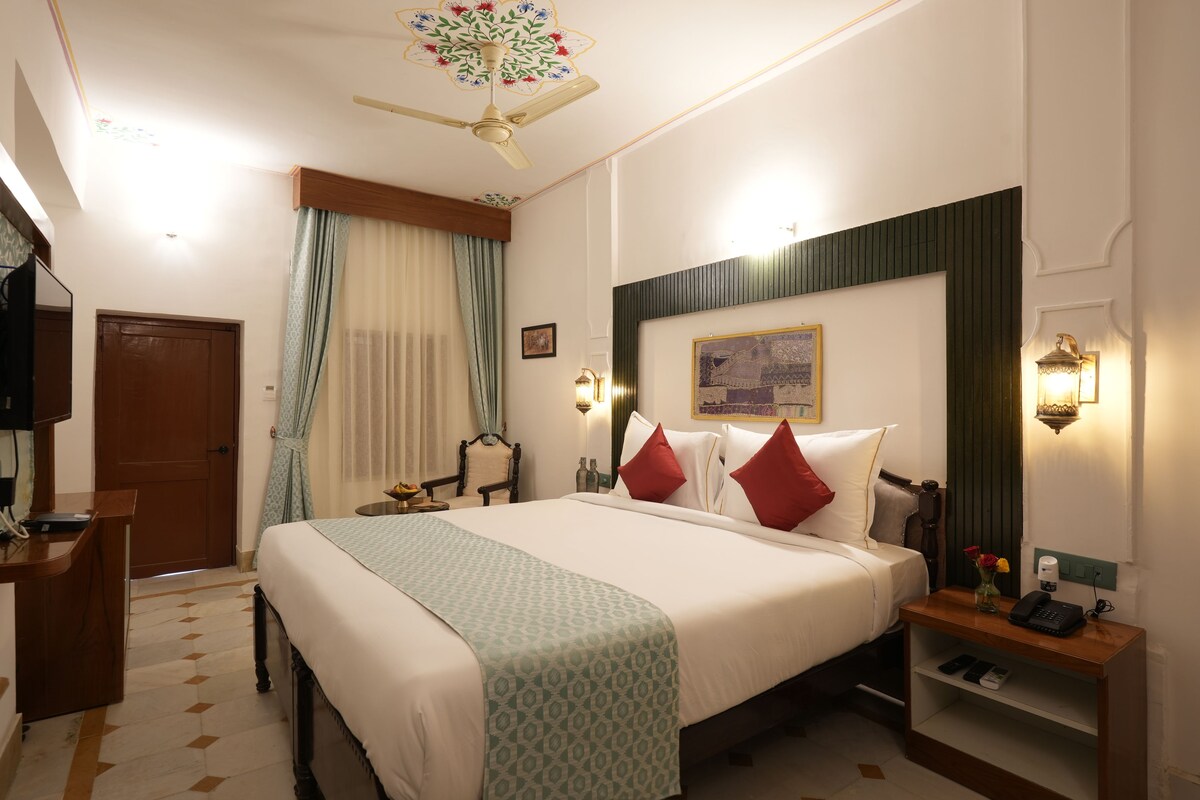 Habitación de hotel con cama, escritorio, silla, televisor, ventilador de techo y ventana con cortinas.