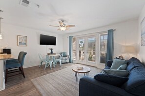 A living room with a blue sofa, a dining area with chairs, a TV mounted on the wall, and a ceiling fan. - Crystal Beach Condo | Walk to the Gulf (Destin)