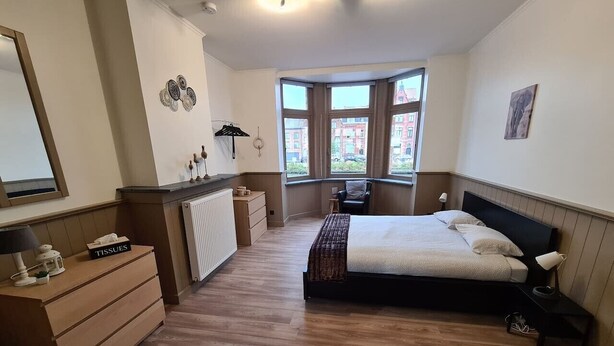Une chambre à coucher comprenant un lit, une commode, un radiateur et une fenêtre donnant sur des immeubles.
