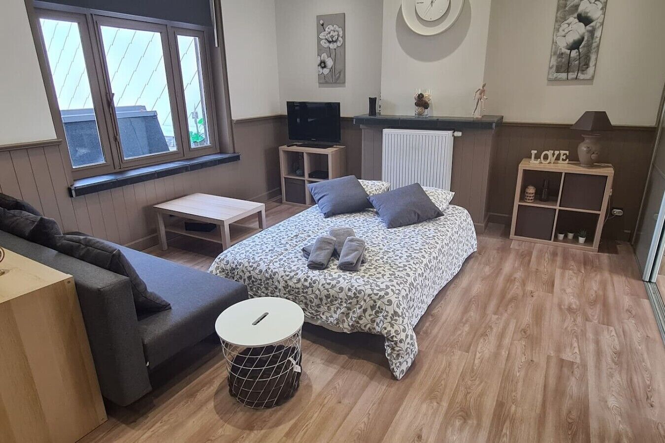 A bedroom with a bed, a TV, a small table, a sofa, a round side table, and a radiator.
