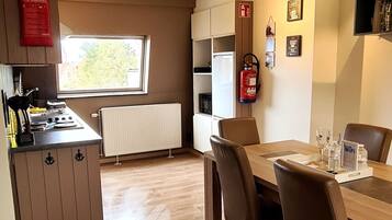 Een kleine keuken met een fornuis, spoelbak en kastjes, een eettafel met stoelen, en een brandblusser aan de muur.