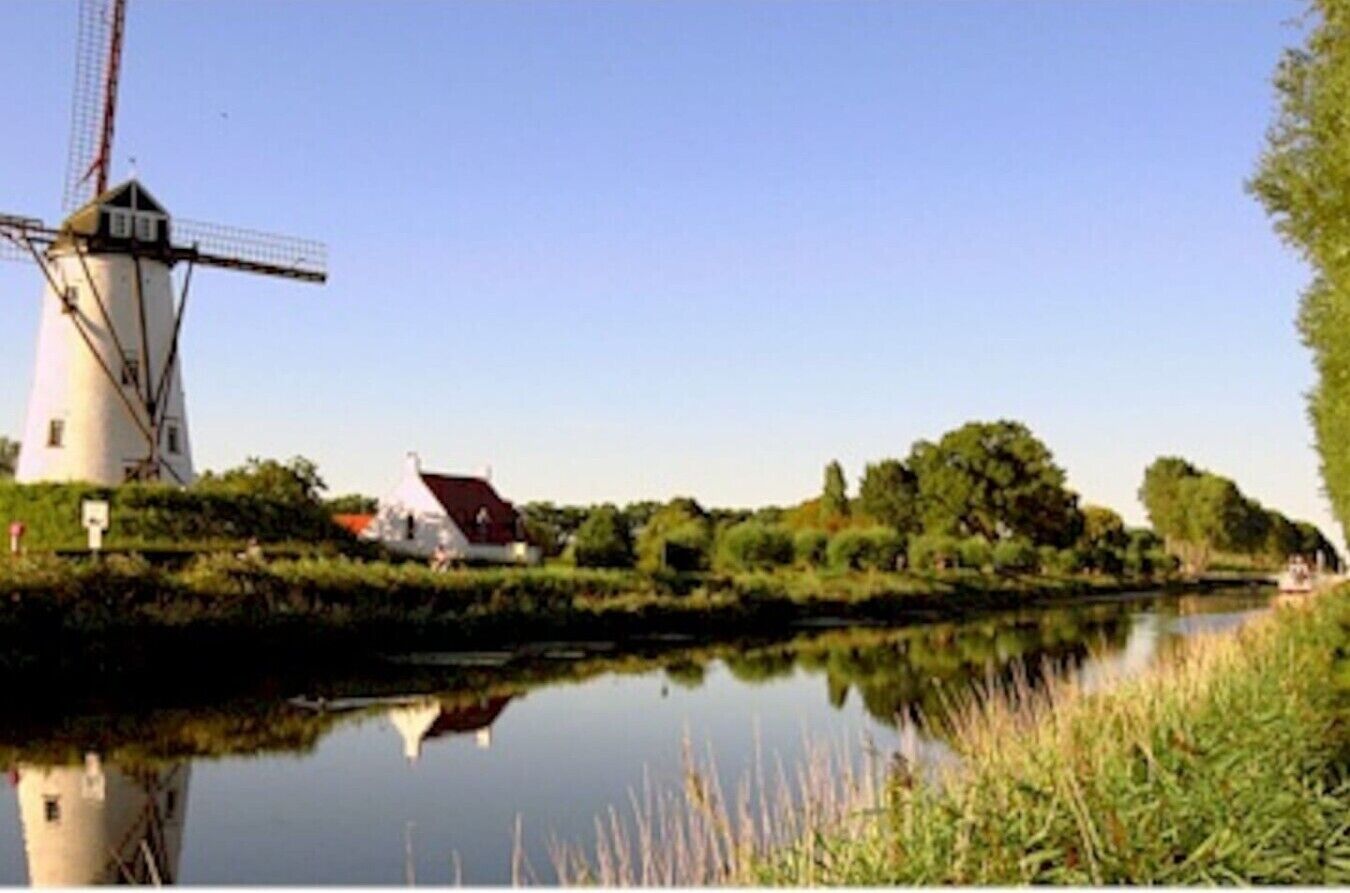 A windmill by a calm river with a house in the background.