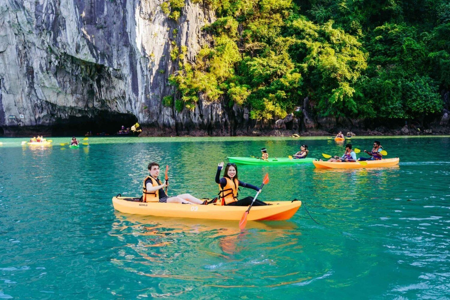 Un grupo de personas en kayaks en aguas tranquilas, con un acantilado rocoso y abundante vegetación al fondo.