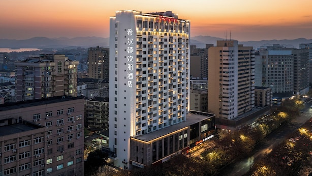 Um hotel moderno com vários andares, uma placa de destaque e vista para o horizonte urbano ao entardecer.