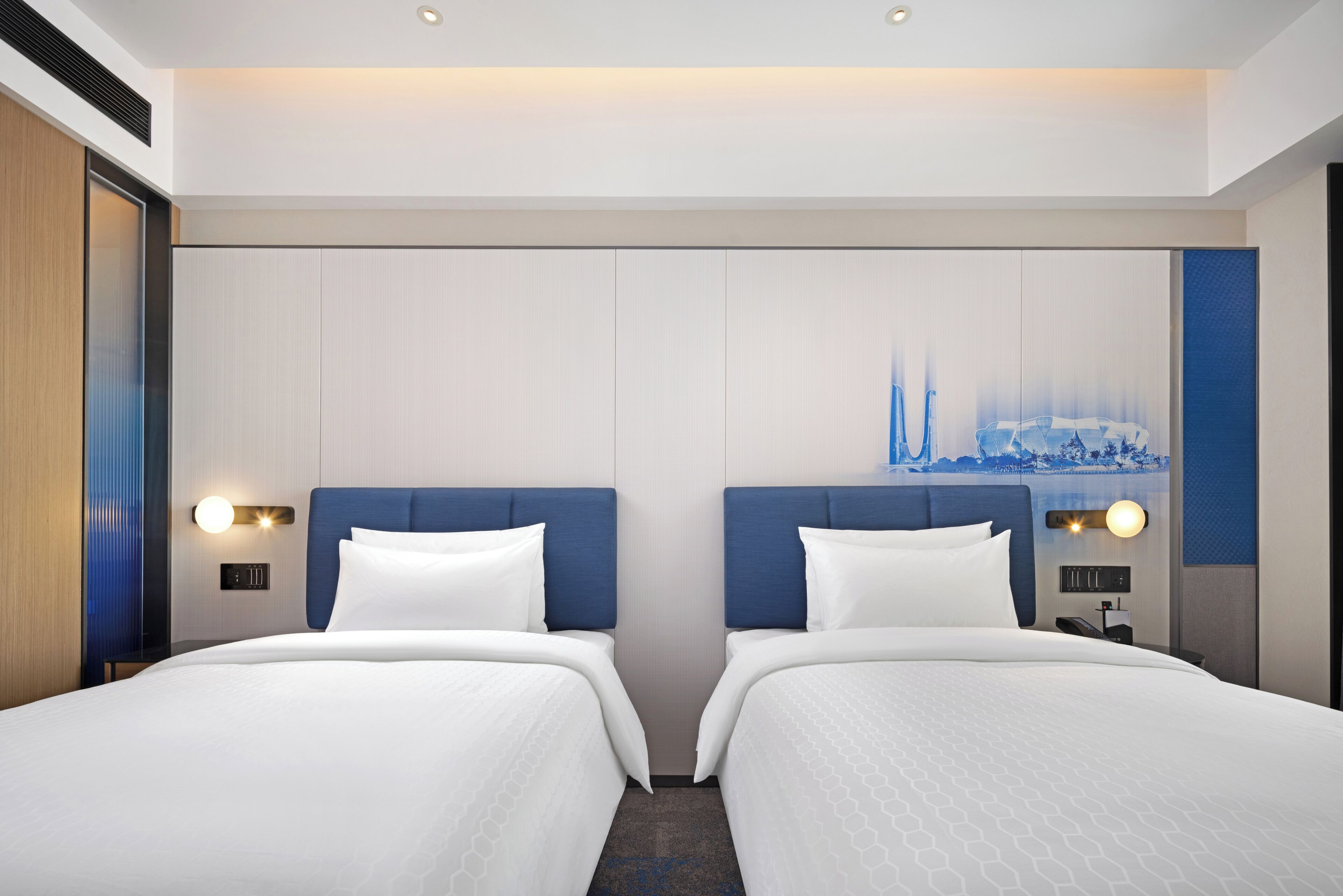 Habitación de hotel con dos camas, un cuadro de paisaje urbano en la pared y un panel de control sobre la mesita de noche.