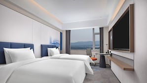 Quarto de hotel com duas camas, uma mesa, uma cadeira, uma TV e uma janela grande com vista para a cidade.