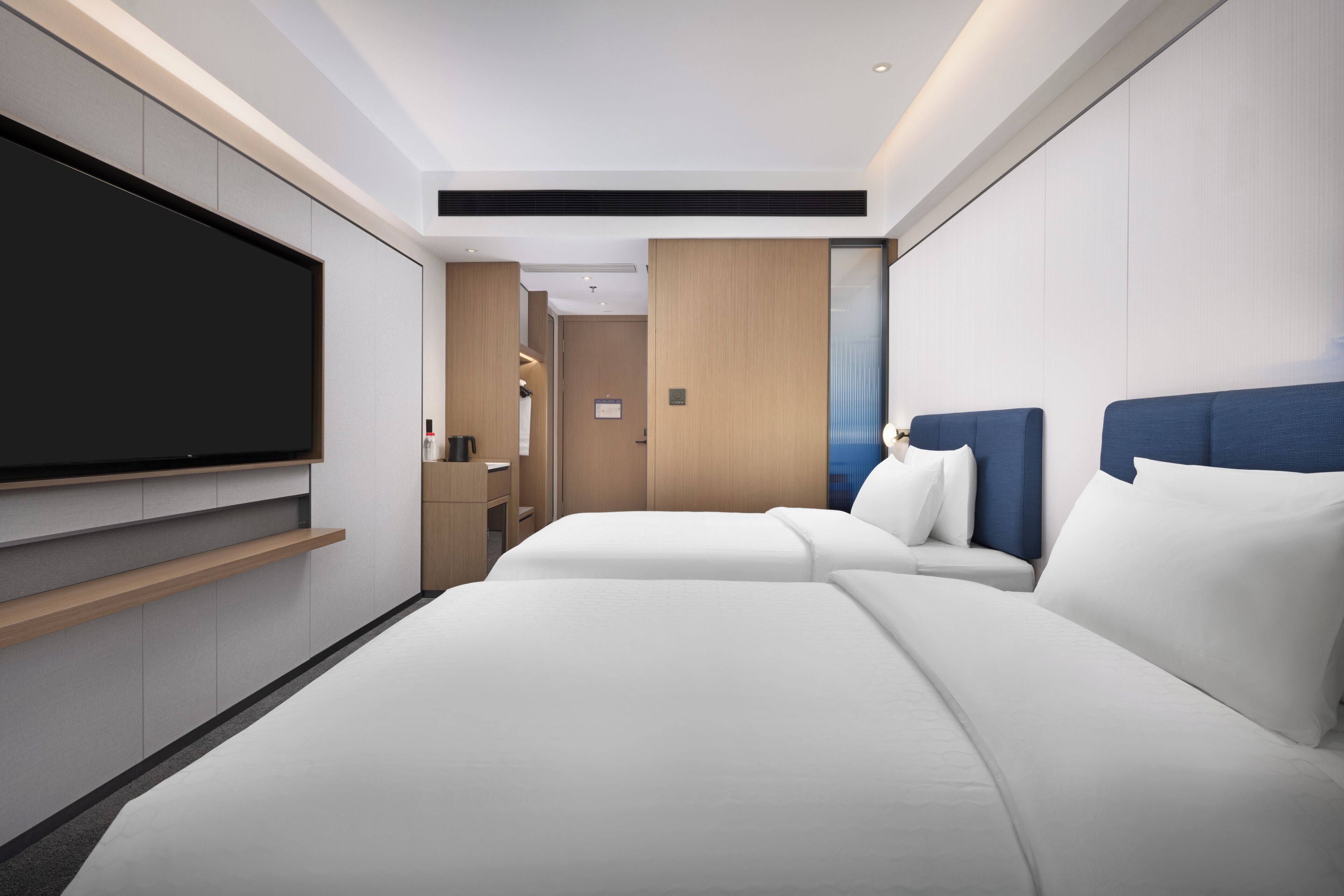 Une chambre d’hôtel moderne dotée d’un grand téléviseur à écran plat fixé au mur, de deux lits avec du linge de lit blanc, d’une tête de lit en bois et d’un petit coin salon avec une table et des chaises.