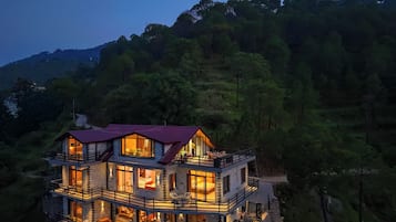 Un edificio de varios pisos con balcones, grandes ventanales y un techo rojo, rodeado de un bosque denso.