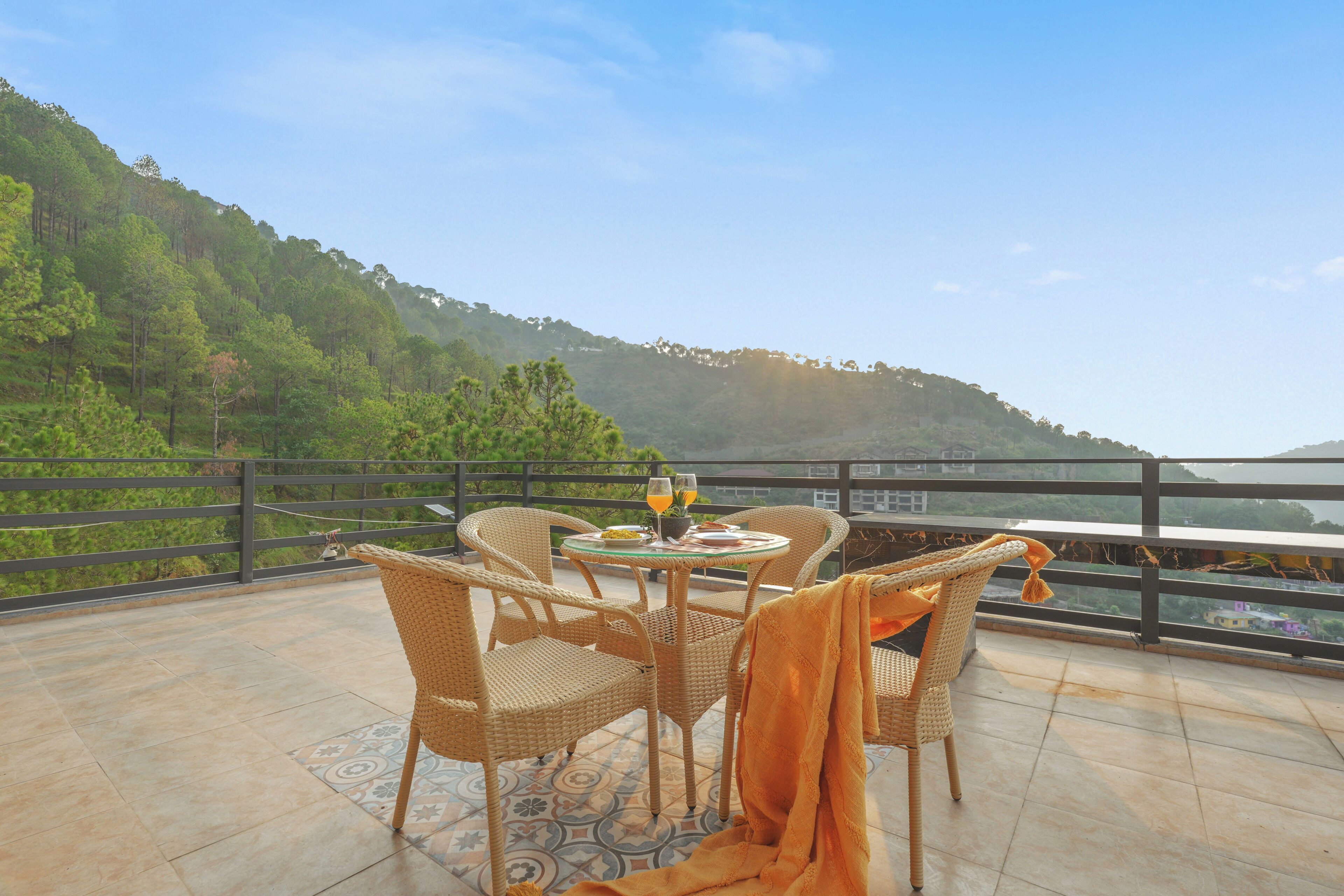 A terrace with wicker furniture, a glass table, and a view of a green hillside.
