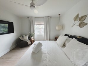 A bedroom with a bed, a ceiling fan, a window with curtains, a bedside table with a plant, and wall-mounted decorative leaves. - The Madonna House | Entire Beach Retreat | 6BD 4BA (St. Augustine)