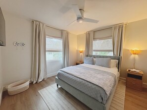 Un dormitorio con una cama, dos mesitas de noche, un ventilador de techo y una ventana con cortinas.