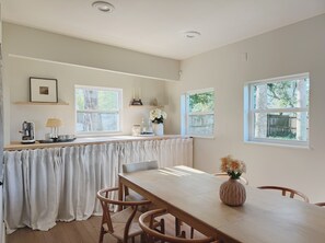 Ein Essbereich mit Tisch, Stühlen und einem Blumenstrauß. Fenster mit Vorhängen, ein Regal mit Dekoration und eine Kaffeemaschine auf der Arbeitsplatte.