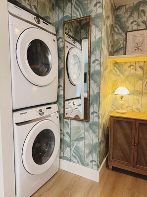 A laundry area with two Samsung washing machines, a mirror, and a wooden cabinet with a lamp. - The Madonna House | Entire Beach Retreat | 6BD 4BA (St. Augustine)