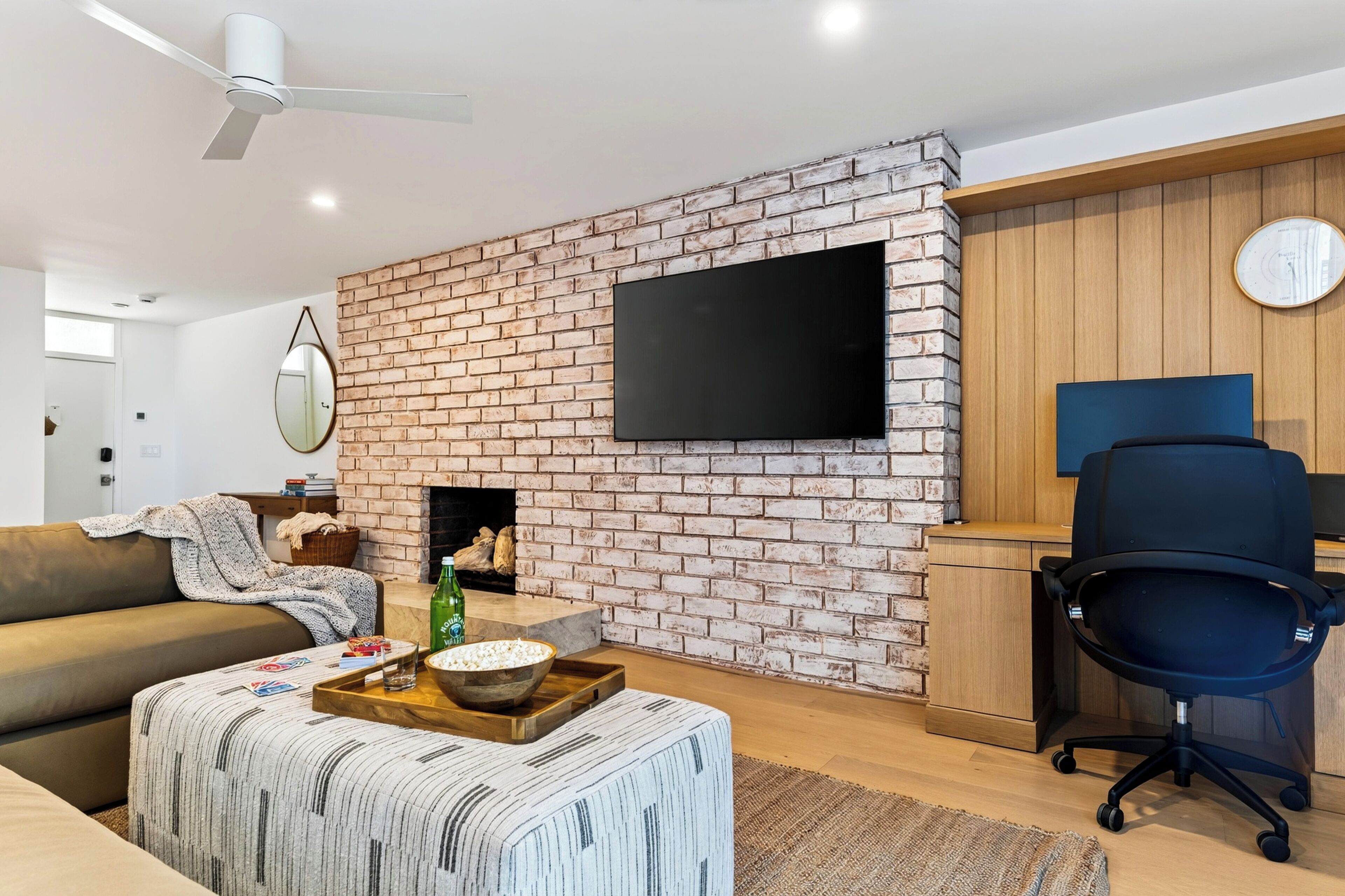 Una sala moderna con pared de ladrillo, televisor de pantalla plana, mueble de madera, silla de oficina azul y un rincón de descanso con sofá y reposapiés.