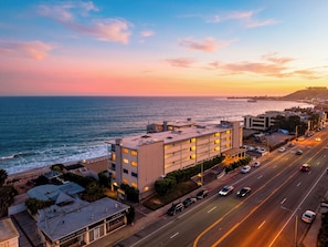 A coastal building with multiple floors, a parking area, and a road with vehicles.