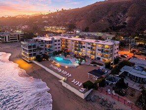 Un hotel frente a la playa con varios pisos, una piscina y una zona de playa con tumbonas.