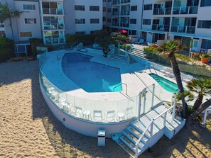 A pool area with a curved design, surrounded by a sandy beach and palm trees.