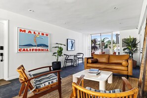 Un salon moderne avec un canapé en cuir marron, des chaises en bois, une table basse et une grande affiche représentant Malibu, en Californie.