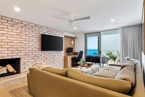 A modern living room with a brick wall, a flat-screen TV, a fireplace, a sofa, a dining area with a table and chairs, and a balcony with a view.