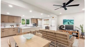 Eine moderne Küche mit Holzausstattung, ein Essbereich mit einer Glas-Schale als Mittelpunkt und ein Wohnbereich mit Sofa und Fernseher.