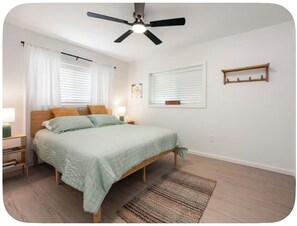 Un dormitorio con una cama, ventilador de techo, mesita de noche con lámpara y ventana con persianas.