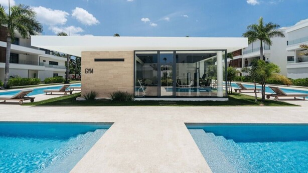 A modern building labeled 'GYM' with a glass entrance, surrounded by a swimming pool area with lounge chairs. - 2BR Ground Floor Beach Retreat | Partial View (Las Terrenas)