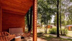A wooden deck with outdoor furniture, a covered seating area, and a view of a forested area with cabins. - Premium Waterfront Scandi Lodge (Wilsum)