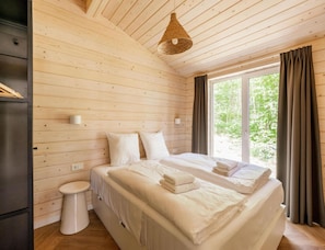 A bedroom with a wooden ceiling, a bed with white linens, a bedside table, a wall-mounted light, and a window with curtains. - Premium Waterfront Scandi Lodge (Wilsum)