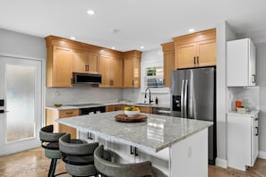 A modern kitchen with wooden cabinets, a stainless steel refrigerator, a marble countertop, and a bowl of fruit. - Baby Be Sweet by AvantStay | Pool + Gazebo Lounge | Walk to Dining (Oakland Park)