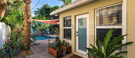 Un jardin arrière d’une maison, avec une piscine, des plantes en pot et un patio couvert.